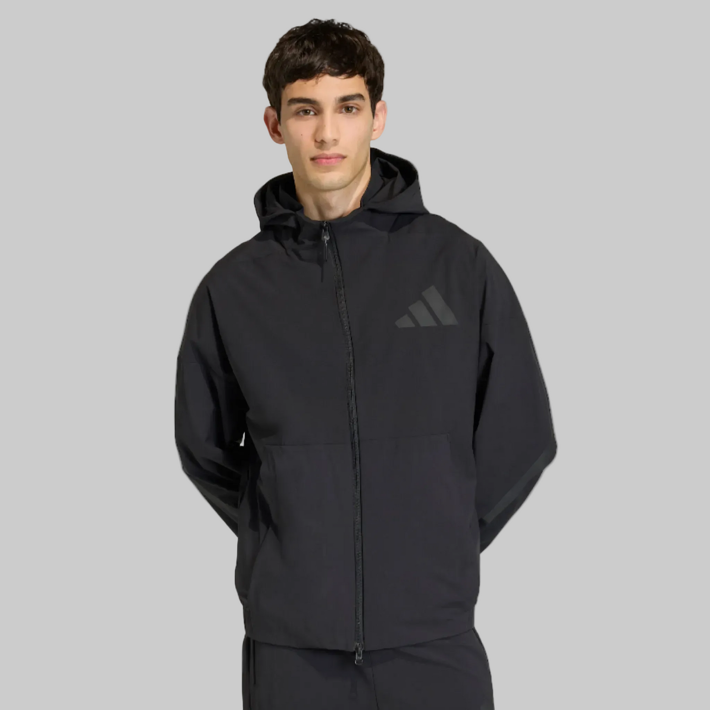 A man wearing a black Adidas hooded jacket with a subtle tonal logo on the chest, photographed against a clean light gray studio background.